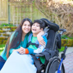 Smiling teenage girl hugging disabled nine year old brother in wheelchair outdoors
